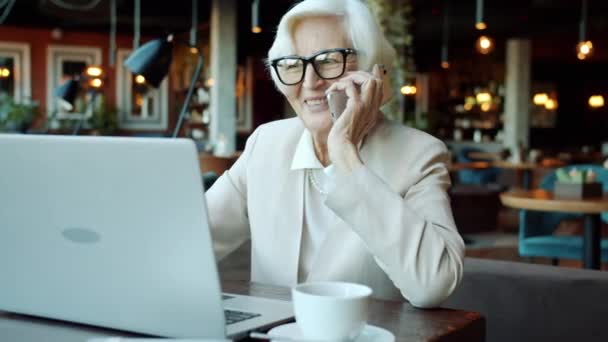 Alegre señora mayor hablando por teléfono móvil y trabajando con el ordenador portátil en la cafetería — Vídeos de Stock