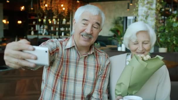 Старший муж и жена используют смартфон, чтобы сделать селфи в ресторане, в то время как женщина держит цветы — стоковое видео