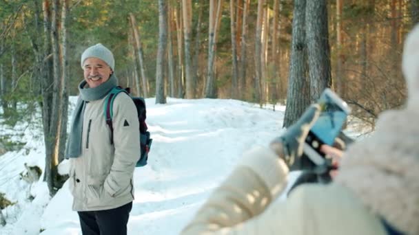 Hombre mayor con mochila posando para la cámara del teléfono inteligente en el parque de invierno, mientras que la mujer toma fotos — Vídeo de stock