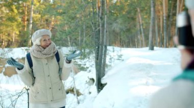 Kocası kışın ormanda kamerayla fotoğraf çekerken kameraya poz veren mutlu yaşlı kadın yürüyüşçü.