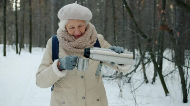 Красивая пожилая женщина пьет чай из термоса во время приятной прогулки в зимнем парке — стоковое видео