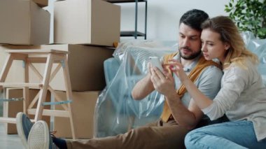 Neşeli kız ve erkek akıllı telefon kullanıyor ekranlara dokunuyor ve yeni dairenin içini işaret ediyor.