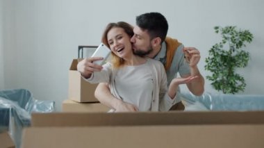 Neşeli kız ve erkek, taşınma sırasında akıllı telefon kamerasıyla yeni ev anahtarlarıyla selfie çekiyorlar.