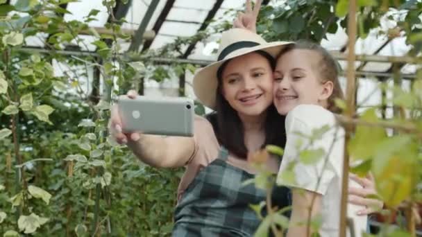 Cámara lenta de madre e hija tomando selfie en invernadero posando besándose sonriente — Vídeos de Stock
