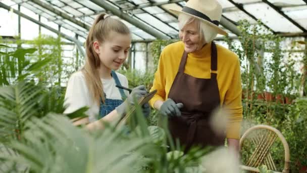 Granny checking plants in orchard and talking while granddaughter taking notes in notebook — Stock Video