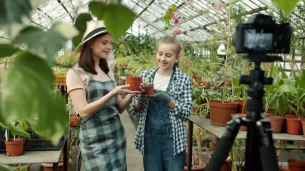 Mother and daughter vloggers recording video in greenhouse waving hand talking showing thumbs-up gesture — Stock Video