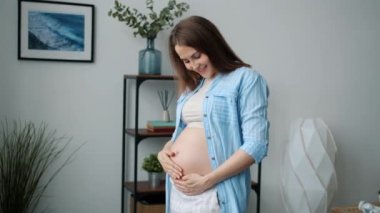 Karnına dokunan ve kameraya bakıp gülümseyen neşeli hamile kadının ağır çekimde portresi.