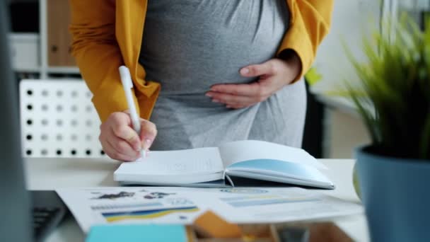 Empregada grávida trabalhando no escritório escrevendo em caderno de pé na mesa — Vídeo de Stock