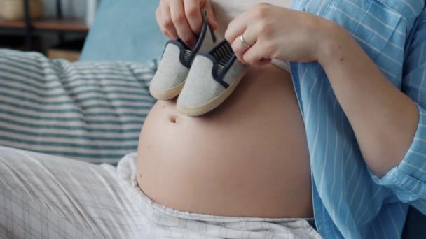 Close-up van zwangere buik en vrouwelijke handen houden baby laarzen en lopen op het lichaam — Stockvideo