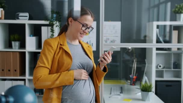 職場でスマートフォンのタッチスクリーンを使用して陽気な期待の母親のオフィスの労働者 — ストック動画