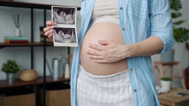 Detailní záběr na roztomilé těhotné břicho a ženy ruku drží ultrazvuk obraz zdravého dítěte v bytě — Stock video