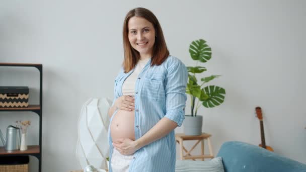 Portrait de belle femme enceinte souriante regardant la caméra toucher le ventre à la maison — Video