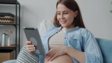 Neşeli hamile kadın tablet ekranına dokunuyor ve gülümsüyor evde teknolojinin tadını çıkarıyor.