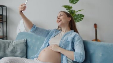 Akıllı telefon kamerasıyla evde poz verip selfie çeken mutlu hamile bir kız.