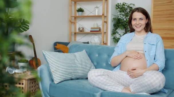 W ciąży młoda kobieta wyrażając miłość do dziecka głaskanie brzuch uśmiechnięty siedzi na kanapie w domu — Wideo stockowe