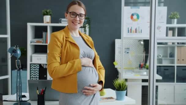 Portrait au ralenti d'une employée enceinte debout dans une pièce de bureau touchant le ventre en souriant — Video