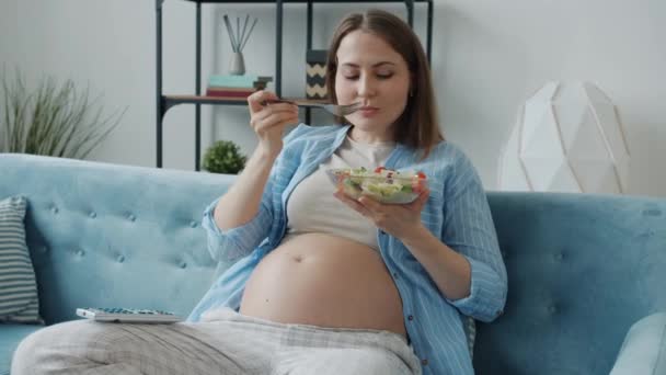 Vidám terhes nő élvezi étkezés friss saláta és tévézni otthon — Stock videók