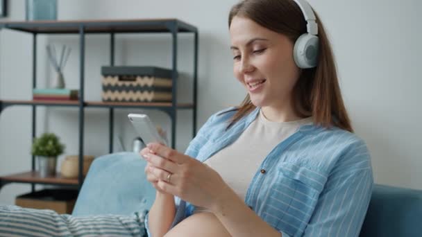 Slow motion of joyful young expecting mother listening to music in headphones and using smartphone — Stock Video