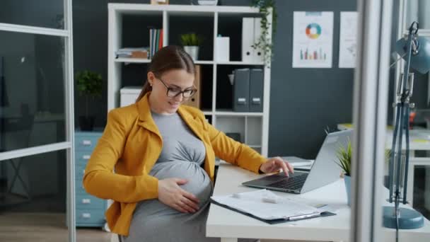 Mulher de negócios grávida alegre usando computador portátil e barriga carinhosa no local de trabalho — Vídeo de Stock