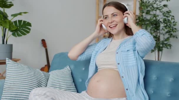 Radosne oczekiwanie matki słuchającej muzyki, a następnie zakładającej słuchawki na brzuch, dzielącej się zabawą z dzieckiem — Wideo stockowe