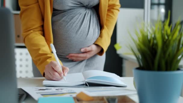 Middle shot of pregnant businesswoman writing in notebook working in office and touching belly — Stock Video