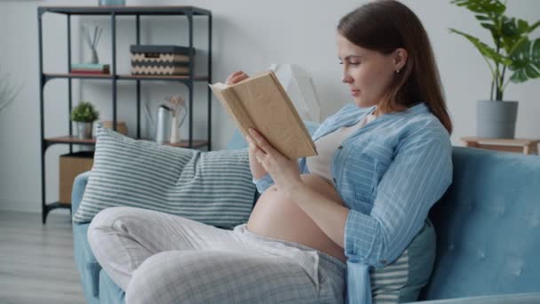 Vacker gravid kvinna läser bok sitter på soffan hemma ler njuter litteratur — Stockvideo