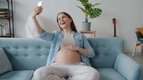 Beautiful expecting mother taking selfie with smartphone camera sitting on sofa at home — Stock Video