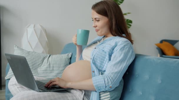 Powolny ruch oczekiwania matki cieszącej się drinkiem i korzystającej z laptopa piszącej uśmiech w domu — Wideo stockowe