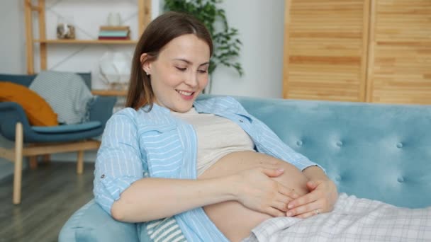 Hidas liike iloinen raskaana oleva nainen hyväilee hänen vatsansa ja hymyilee rentouttava sohvalla kotona — kuvapankkivideo