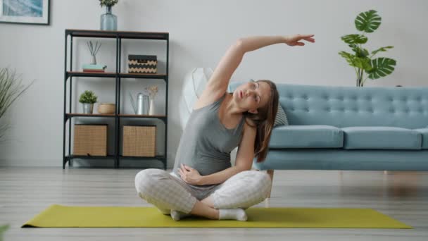 Медленное движение привлекательной беременной женщины занимающейся спортом дома растягивая тело сидя на коврике для йоги — стоковое видео