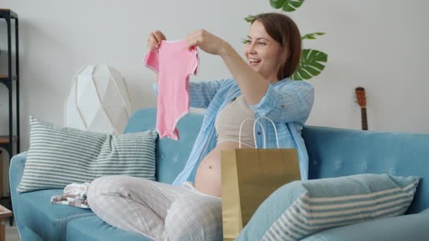 Movimiento lento de la mujer embarazada joven tomando ropa de bebé diminuta de la bolsa de compras en el sofá en casa — Vídeo de stock