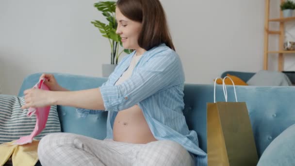 Långsam rörelse av leende gravid kvinna njuter av små baby kläder håller på magen — Stockvideo