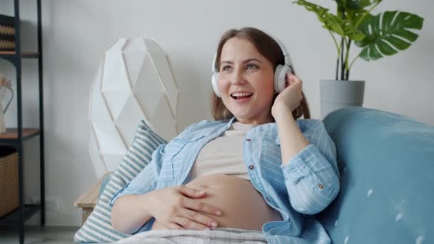 Cámara lenta de la madre embarazada disfrutando de la música en los auriculares cantando y divirtiéndose en casa — Vídeos de Stock