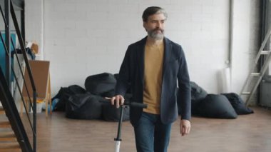 Olgun bir adam mobiletiyle ofise geliyor ve el sallayarak gülümsüyor.