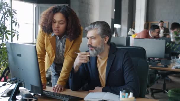 Dojrzałe biały mężczyzna i młody Afroamerykanka współpracownicy mówi patrząc na ekranie komputera — Wideo stockowe