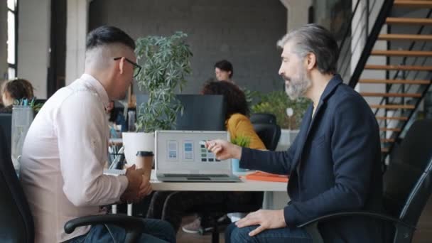 Коллеги-мужчины говорят о приложениях для смартфонов, которые смотрят на экран ноутбука на рабочем месте — стоковое видео