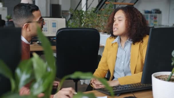 Hombre árabe y mujer afroamericana hablando y riendo en el escritorio en la oficina — Vídeos de Stock