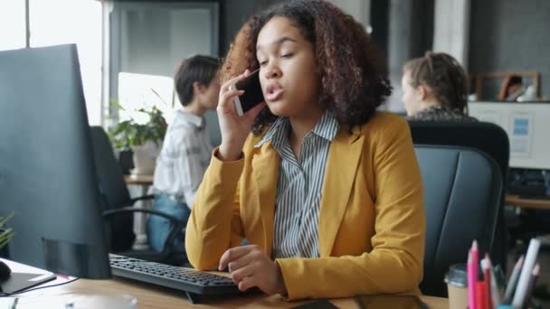 Радостная афроамериканка, болтающая по мобильному телефону и пользующаяся компьютером в современном офисе — стоковое видео