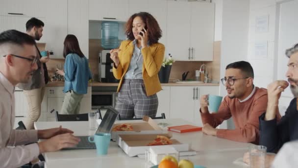 Afro-americká žena mluví na mobilním telefonu jíst pizzu, zatímco kolegové mluví těší oběd v práci — Stock video