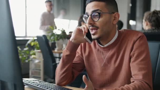 Ernstige Midden-Oosten man belt op mobiele telefoon en kijkt naar het computerscherm in het kantoor — Stockvideo