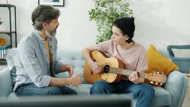 Турбота тато навчає сина підлітка, як грати на гітарі розмовляючи і жестикулюючи в приміщенні вдома — стокове відео