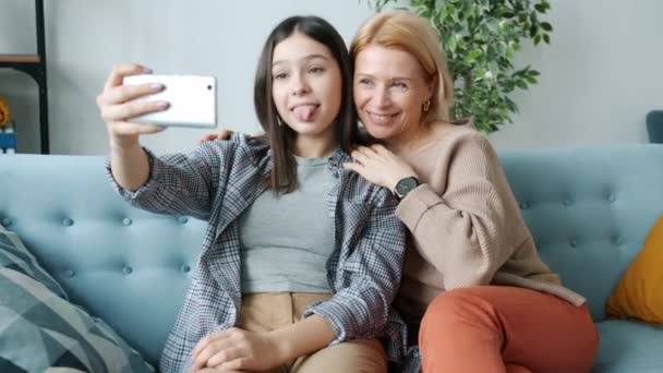 Девочка-подросток делает селфи с мамой, развлекаясь, делая смешные лица, используя камеру смартфона — стоковое видео
