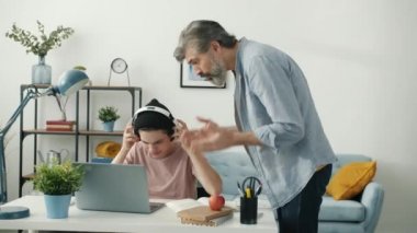 Kulaklıklı çocuk, kızgın babası gelip ödevlerini yaparken dizüstü bilgisayar kullanıyor.