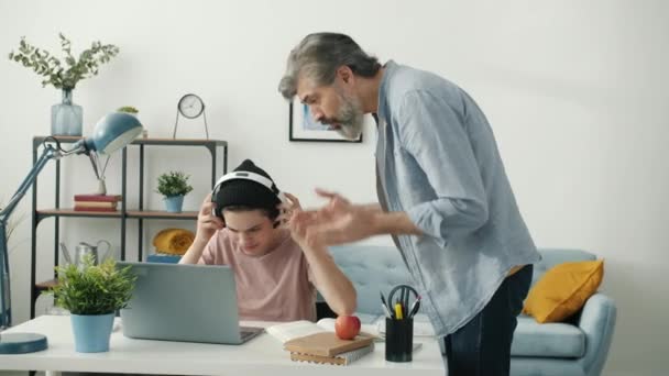 Kulaklıklı çocuk, kızgın babası gelip ödevlerini yaparken dizüstü bilgisayar kullanıyor. — Stok video