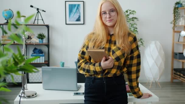 Повільний рух розумної дівчини-підлітка, що стоїть вдома, тримає книгу посміхаючись дивитися на камеру — стокове відео