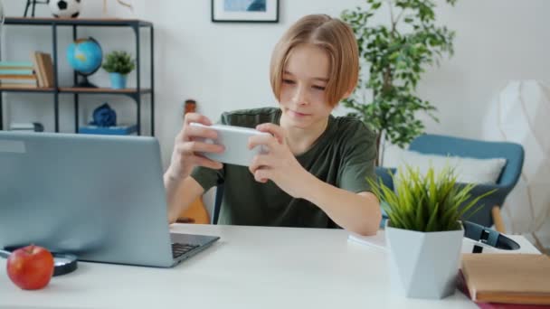 Усміхнена дитина-підліток насолоджується відеоіграми, торкаючись екрану смартфона, розважаючись вдома — стокове відео