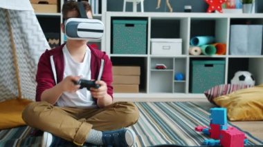 Oyun oynayan çocuk, evde yerde oturmuş joystick ve sanal gerçeklik gözlüğü kullanıyor.