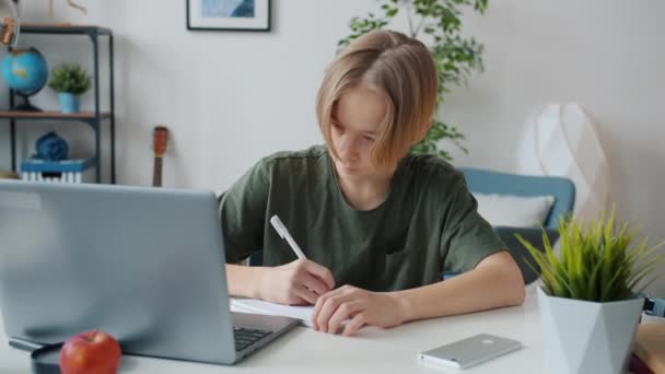 Хлопчик-підліток пише в блокноті і працює з ноутбуком, роблячи домашнє завдання в квартирі — стокове відео
