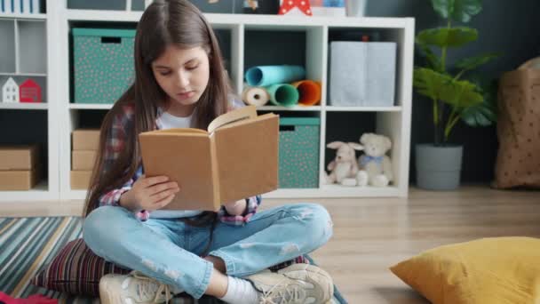 Chica seria leyendo libro sentado en el suelo en casa concentrado en la actividad intelectual — Vídeos de Stock