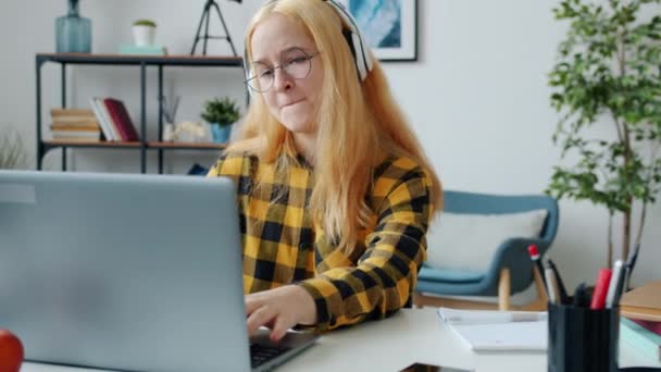 Повільний рух радісного підлітка за допомогою ноутбука друку та прослуховування музики в навушниках — стокове відео